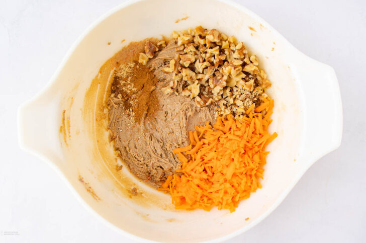 A white mixing bowl containing partially mixed brown batter, chopped walnuts, and shredded carrots, ready to be combined.