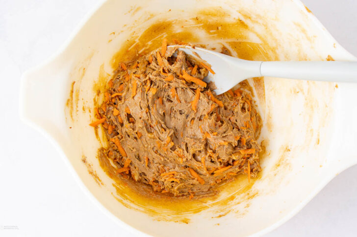 A white mixing bowl filled with thick, orange-brown carrot cake batter. A gray spatula is resting inside, with some batter clinging to it. Shredded carrots and spices are visible in the mixture.