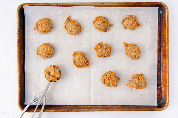 A baking sheet lined with parchment paper holds eleven dollops of cookie dough. A cookie scoop with more dough is positioned on the lower left of the tray, ready to drop another portion.
