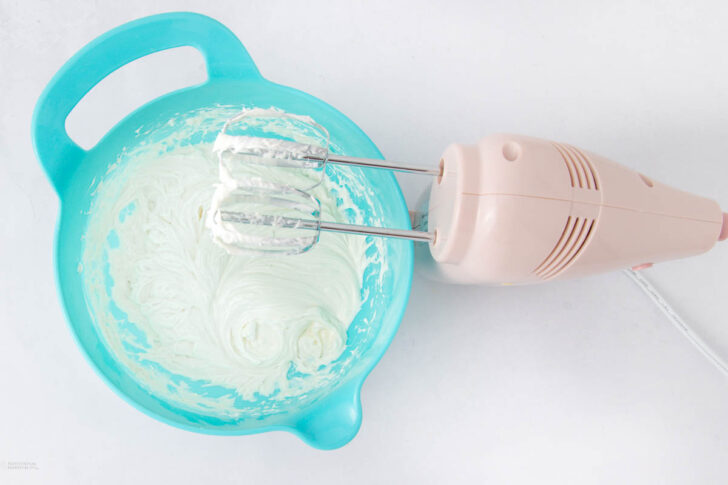 A hand mixer with beaters is resting in a turquoise mixing bowl filled with creamy white frosting. The scene is set on a white surface.