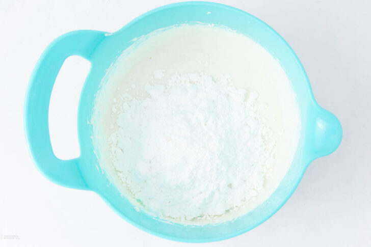 A light blue mixing bowl contains a mound of white powdered sugar on top of a creamy mixture, viewed from above on a white surface.