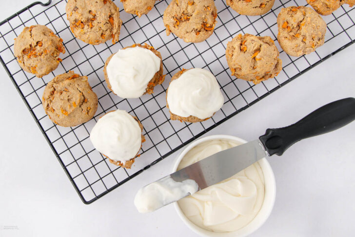 Cookies cooling on a wire rack, some topped with white frosting. A bowl of frosting and a spreading knife with frosting on it are nearby on a white surface.