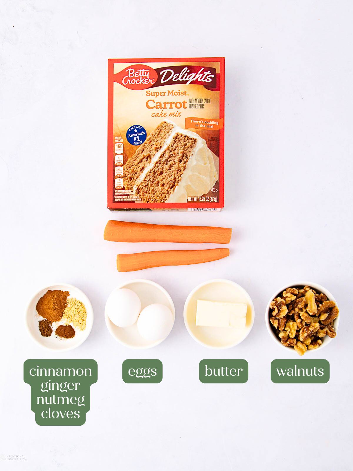 A box of carrot cake mix, two whole carrots, small bowls holding spices (cinnamon, ginger, nutmeg, cloves), two eggs, butter, and walnuts are arranged neatly on a white background.