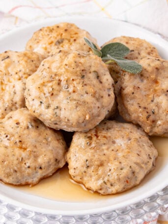 A white plate of six golden-brown turkey breakfast sausage patties garnished with fresh sage, sitting in a small pool of syrup. The plate is on a glass charger atop a white tablecloth with a pastel plaid pattern.