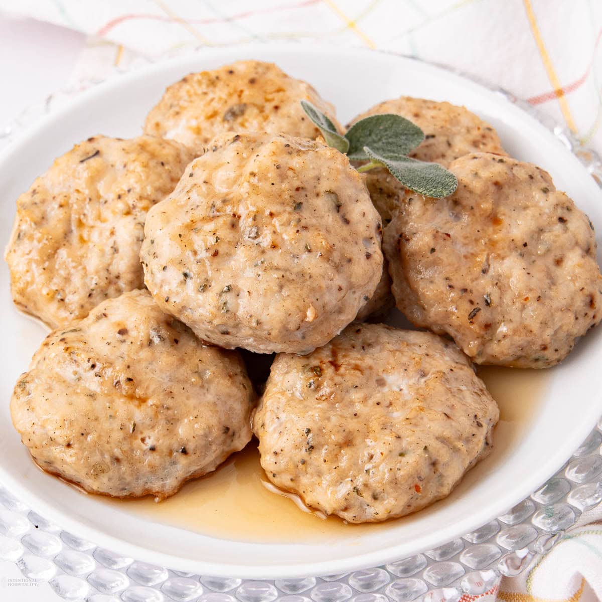 A white plate of six golden-brown turkey breakfast sausage patties garnished with fresh sage