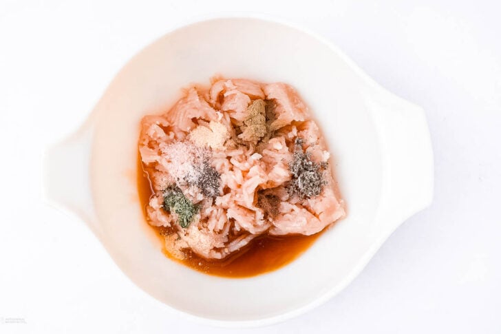 Raw, sliced chicken with various powdered spices and soy sauce is placed in a white mixing bowl on a white background, ready to be marinated.