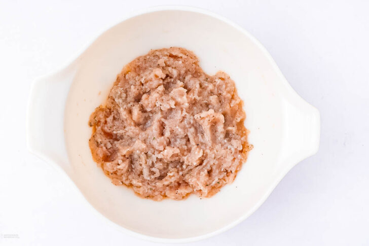 A white bowl containing raw ground chicken on a white background.