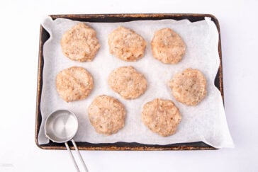 A baking tray lined with parchment paper holds eight raw, round chicken patties. A metal measuring cup rests on the tray’s bottom left corner. The background is a plain white surface.