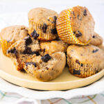 A plate of chocolate chip muffins, some whole and some broken in half, revealing chunks of chocolate inside. The muffins are in yellow-striped paper liners and stacked on a light-colored plate.