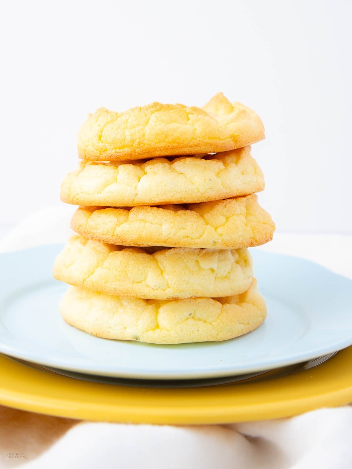 A stack of five golden, fluffy cookies is arranged on a light blue plate, which sits on top of a yellow plate against a white background.