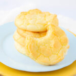 A stack of light, fluffy yellow cookies on a pale blue plate, with one cookie in front showing a large bite taken out of it. The background is softly blurred and bright.