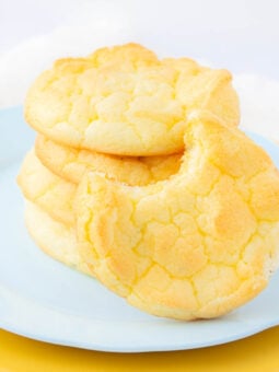 A stack of light, fluffy yellow cookies on a pale blue plate, with one cookie in front showing a large bite taken out of it. The background is softly blurred and bright.