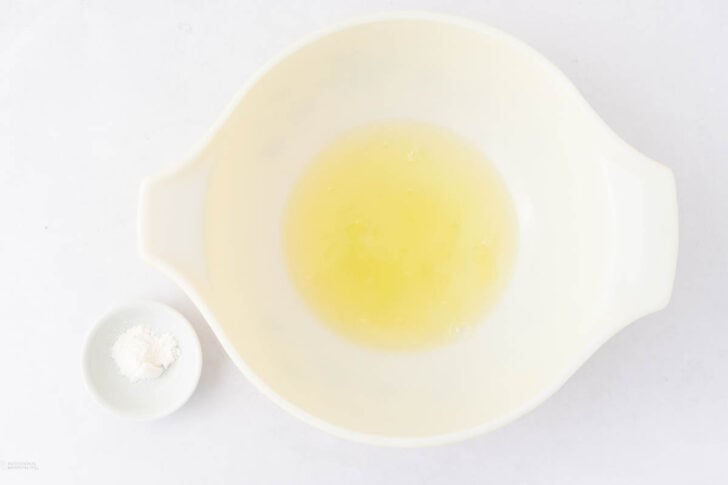 A white mixing bowl containing egg whites sits on a white surface next to a small dish with a small amount of white powder, likely cream of tartar.