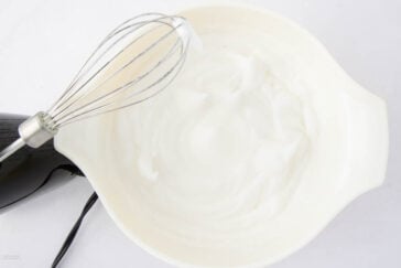 A hand mixer with a whisk attachment is placed over a white bowl filled with stiffly beaten egg whites on a white surface.