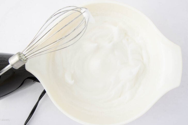 A hand mixer with a whisk attachment is placed over a white bowl filled with stiffly beaten egg whites on a white surface.