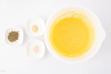 A white bowl with a beaten yellow egg mixture sits beside three small dishes containing dried herbs, garlic powder, and salt on a white surface.