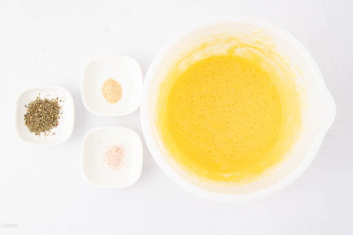 A white bowl with a beaten yellow egg mixture sits beside three small dishes containing dried herbs, garlic powder, and salt on a white surface.