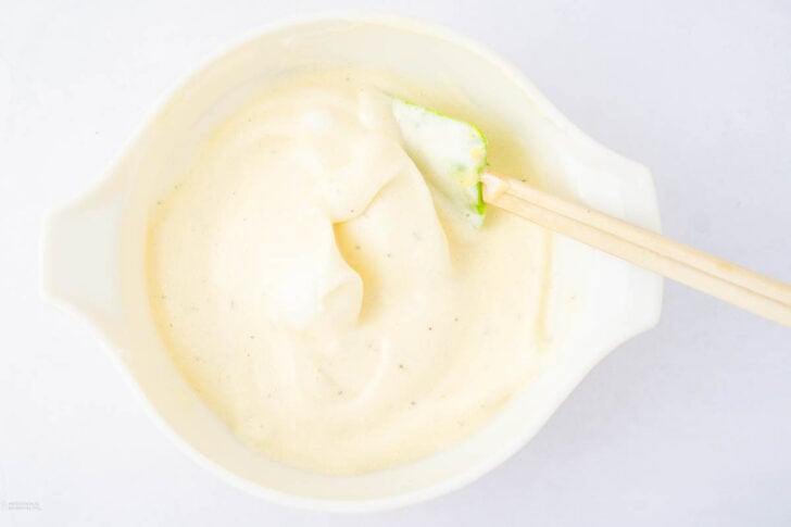 A white mixing bowl filled with a creamy, whipped mixture and a green spatula resting inside, photographed from above on a white surface.