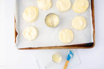 A baking tray lined with parchment paper holds eight unbaked round pastries topped with cream. A metal biscuit cutter sits among them. Below, a spatula and a measuring cup with cream rest on a white cutting board.