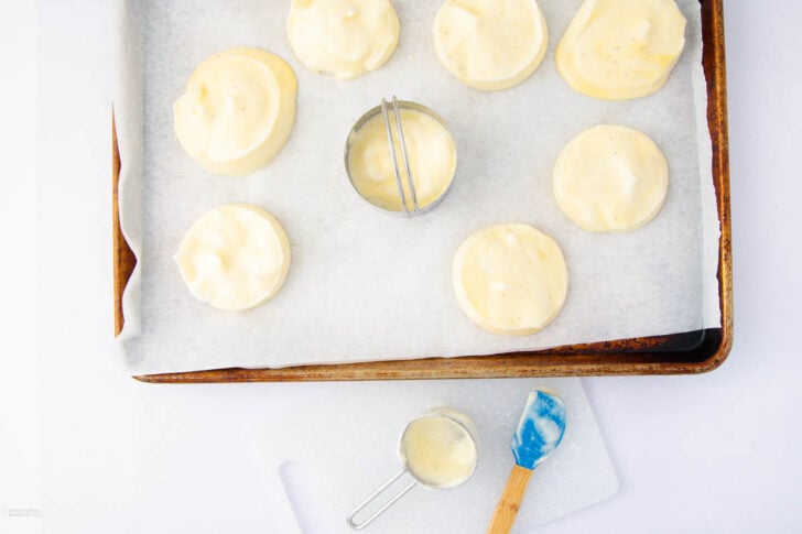 A baking tray lined with parchment paper holds eight unbaked round pastries topped with cream. A metal biscuit cutter sits among them. Below, a spatula and a measuring cup with cream rest on a white cutting board.