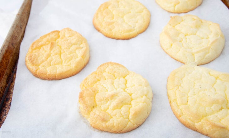 Six golden, slightly cracked cloud bread rounds cooling on a parchment-lined baking sheet. The texture appears light and airy, with a soft, pale-yellow color.