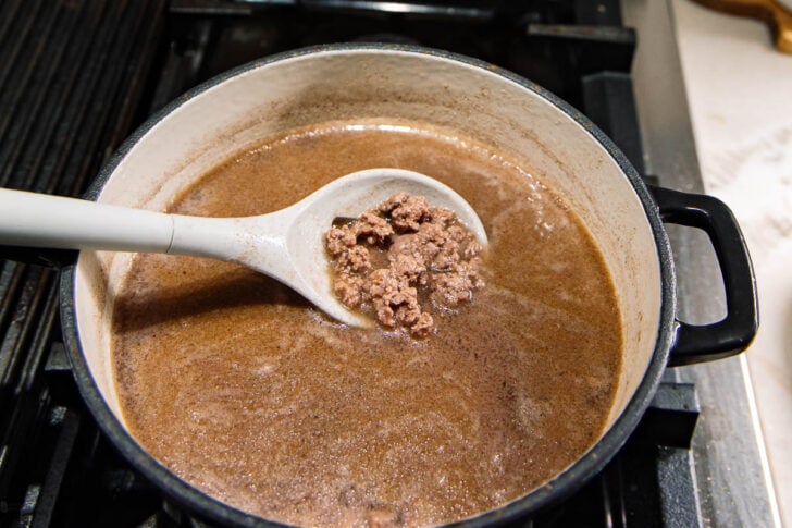 A white ladle holds cooked ground beef above a pot filled with brown, simmering liquid on a stove.