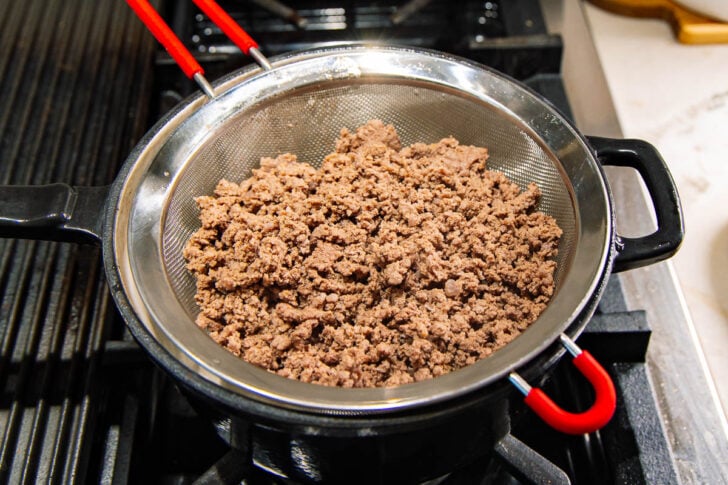 Cooked ground beef is draining in a metal strainer placed over a black pot on a stovetop. Red-handled tongs rest on the edge of the strainer. The kitchen counter and part of the stove are visible.
