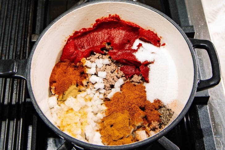 Overhead view of a pot on a stove containing ground meat, chopped onions, tomato paste, sugar, and a variety of colorful spices, all unmixed and ready to be cooked together.