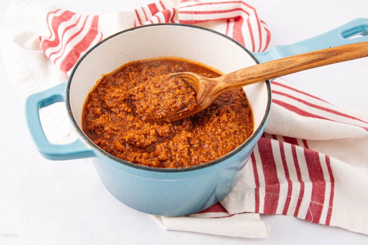 A blue pot filled with thick, hearty chili sits on a red and white striped towel. A wooden spoon rests inside the pot, coated with chili. The background is bright and clean.