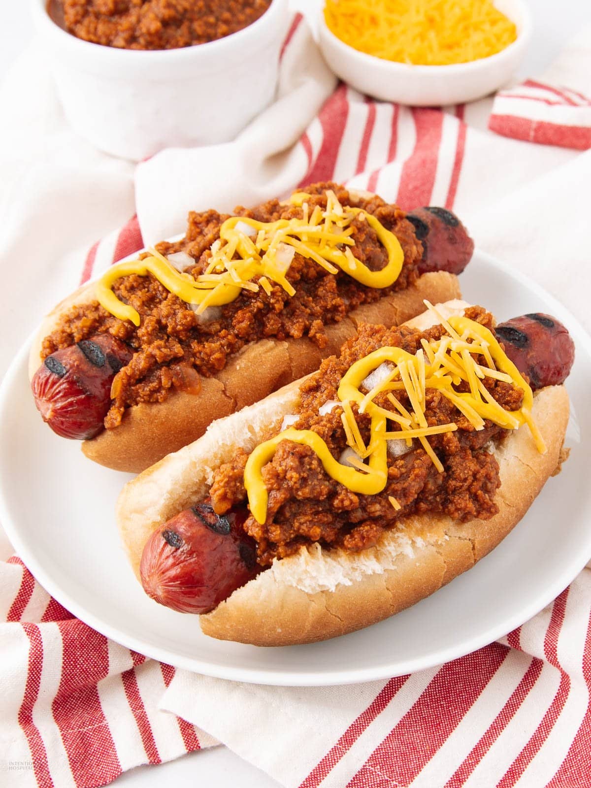 Two grilled hot dogs in buns topped with chili, shredded cheese, and yellow mustard, served on a white plate with a red and white striped napkin underneath. Bowls of shredded cheese and chili are in the background.