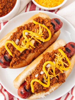 Two grilled hot dogs in buns topped with chili, shredded cheddar cheese, diced onions, and zigzags of yellow mustard, served on a white plate with a red-striped napkin and bowls of chili and cheese nearby.