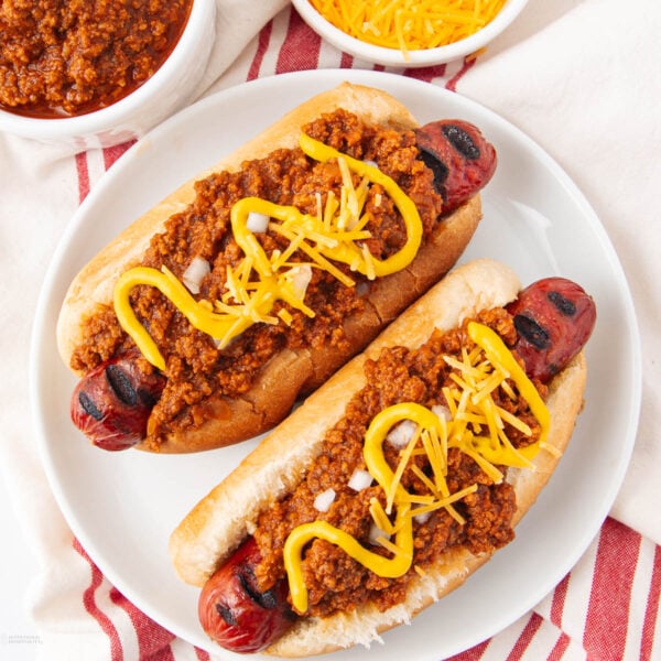 Two grilled hot dogs in buns topped with chili, shredded cheddar cheese, diced onions, and zigzags of yellow mustard, served on a white plate with a red-striped napkin and bowls of chili and cheese nearby.
