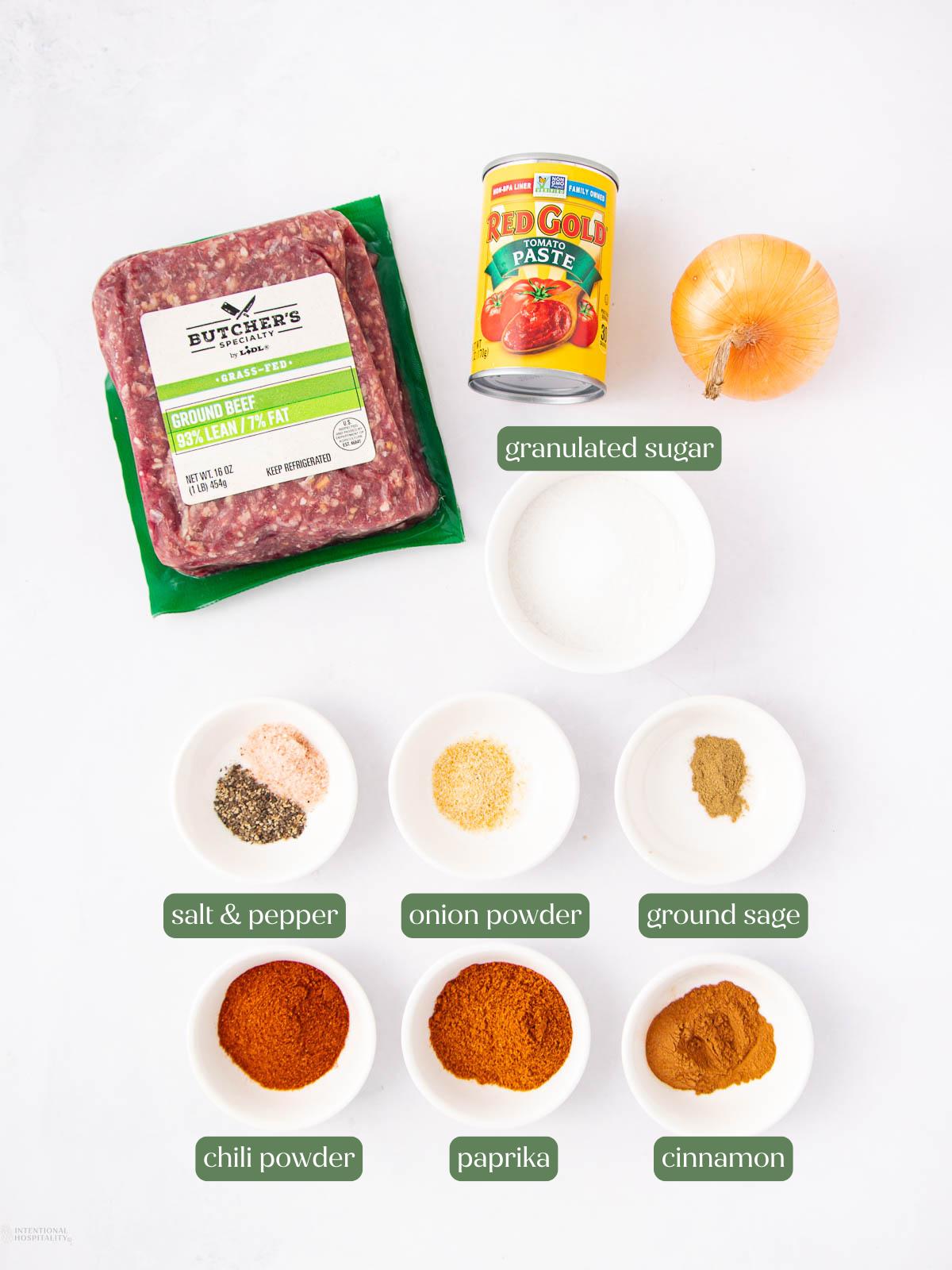 Ground beef, a can of tomato paste, a yellow onion, a bowl of granulated sugar, and small bowls containing salt and pepper, onion powder, ground sage, chili powder, paprika, and cinnamon are arranged on a white surface.
