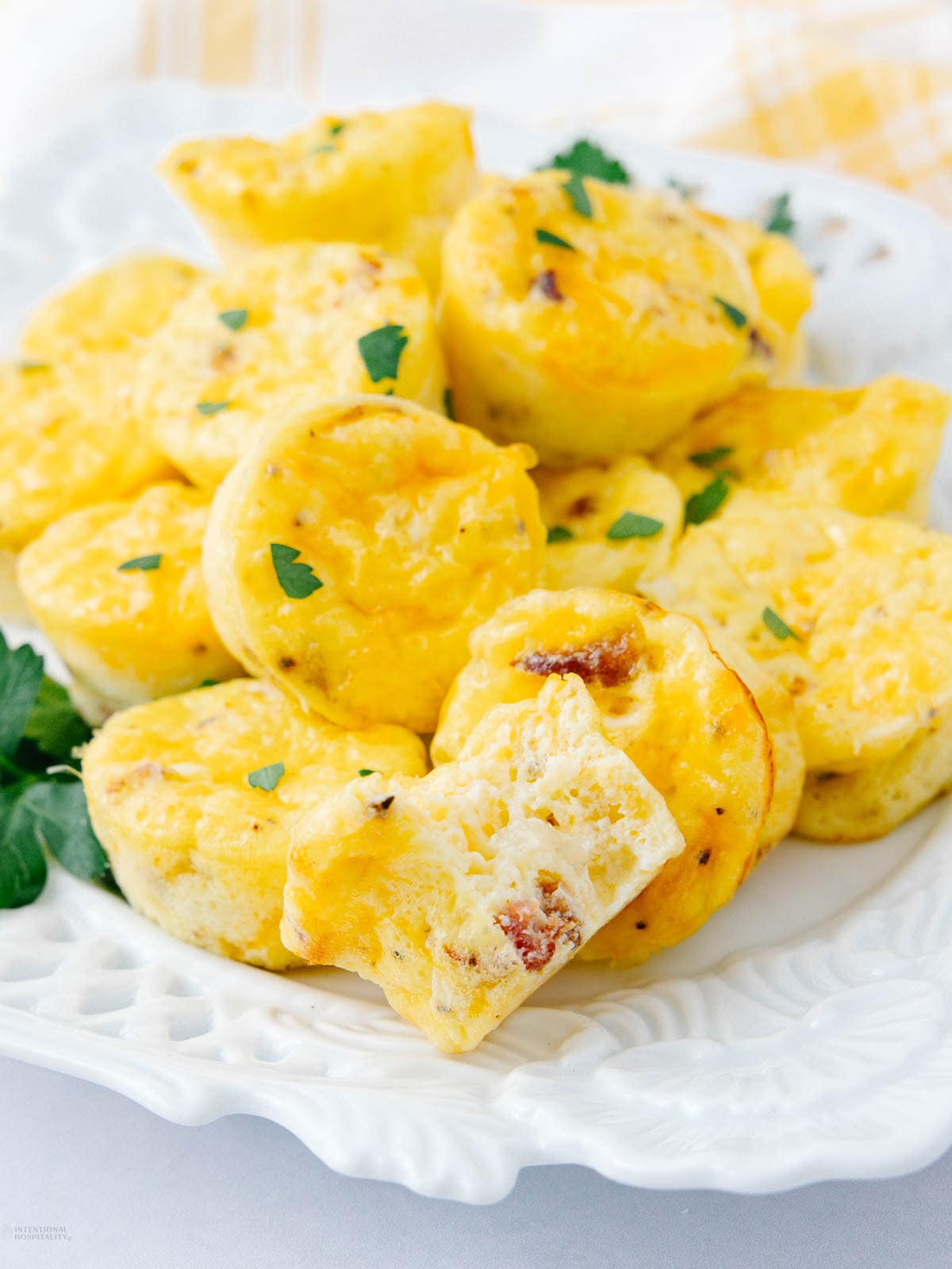 A plate of mini egg muffins garnished with chopped parsley. One muffin in front is cut in half, showing a fluffy inside with pieces of bacon or sausage. The plate is white and serves as a breakfast dish.