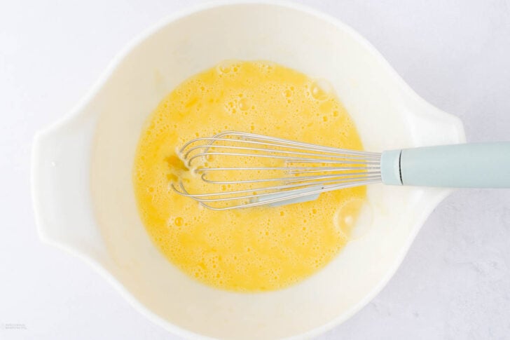A metal whisk with a blue handle rests in a white mixing bowl filled with beaten eggs on a light-colored surface.