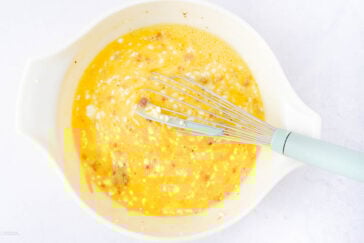 A white mixing bowl contains beaten eggs with visible bits of cheese and meat, being whisked with a light blue-handled whisk on a white surface.