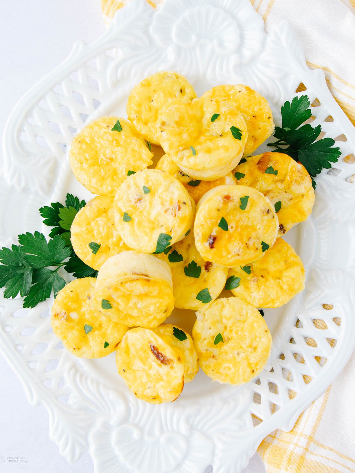 Mini egg muffins garnished with chopped parsley are arranged in a circular pattern on a decorative white plate, with sprigs of fresh parsley placed as garnish on the side.