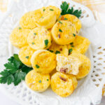 A white plate filled with mini egg muffins garnished with chopped parsley, with one muffin cut open to show the inside. A few sprigs of parsley decorate the plate.