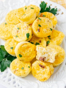 A white plate filled with mini egg muffins garnished with chopped parsley, with one muffin cut open to show the inside. A few sprigs of parsley decorate the plate.