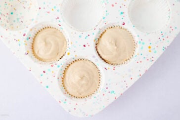 A close-up of a muffin tin with four paper liners, three filled with light tan batter and one empty, on a white surface. The tin is decorated with colorful confetti dots.