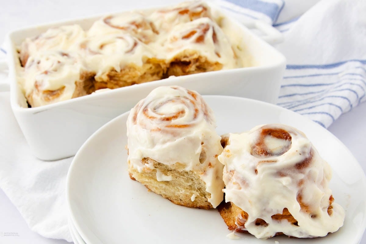 Two iced cinnamon rolls on a white plate, with more cinnamon rolls in a baking dish in the background. All rolls are topped with creamy white frosting. A white and blue striped towel is nearby.