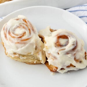 Two cinnamon rolls topped with creamy white icing sit on a white plate, with a hint of a blue and white striped cloth in the background.
