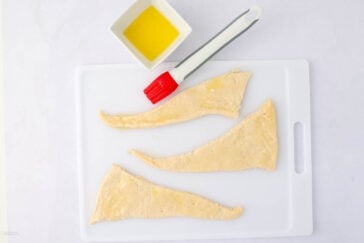 Three unbaked crescent roll dough triangles on a white cutting board, next to a small white bowl filled with melted butter and a red and white pastry brush.