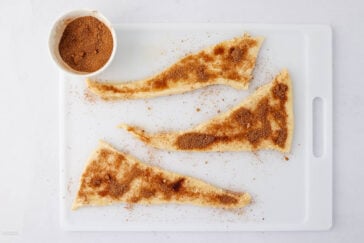 Three uncooked crescent dough triangles topped with cinnamon sugar are on a white cutting board, with a small bowl of cinnamon sugar nearby.