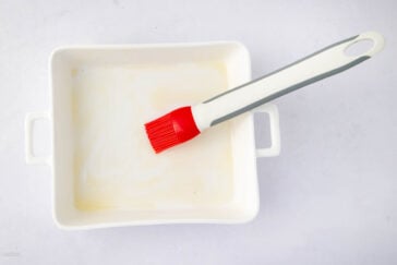 A white rectangular baking dish with handles contains a thin layer of melted butter. A red silicone pastry brush with a white and gray handle rests inside the dish.