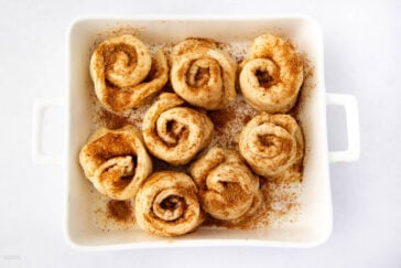 Eight unbaked cinnamon rolls topped and surrounded by cinnamon and sugar are arranged in a white rectangular baking dish on a white background.