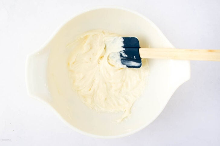 A white mixing bowl containing creamy, whipped mixture being stirred with a blue and wooden spatula, set against a white background.