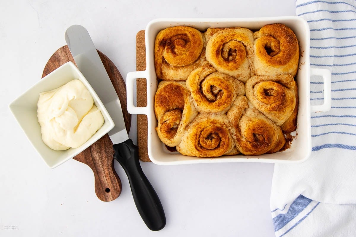 A white baking dish of golden brown cinnamon rolls sits on a table next to a bowl of creamy icing with a spatula, a wooden board, and a white and blue striped kitchen towel.