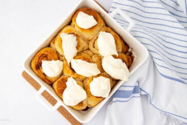 A white baking dish filled with cinnamon rolls, each topped with a generous dollop of cream cheese frosting, sits on a cork mat next to a blue and white striped kitchen towel.