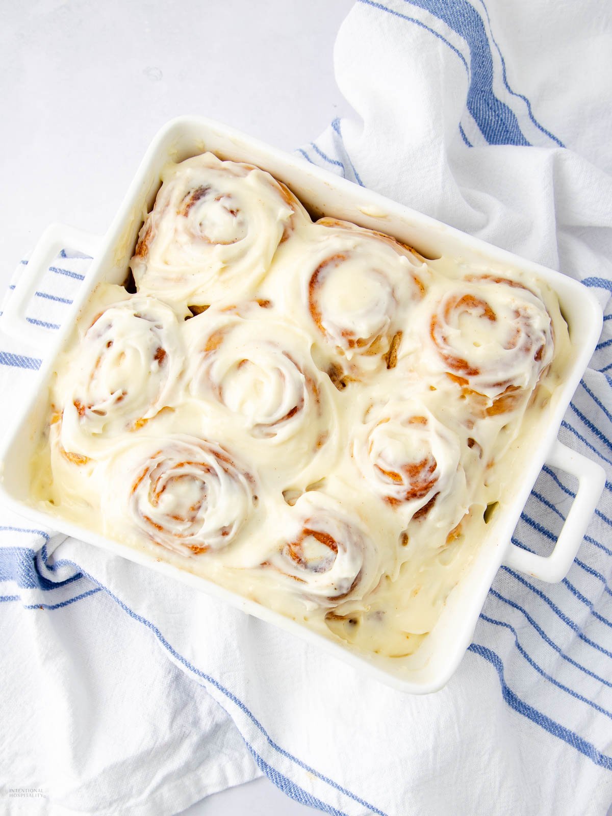 A white baking dish filled with nine cinnamon rolls covered in creamy white icing, sitting on a white and blue striped kitchen towel.
